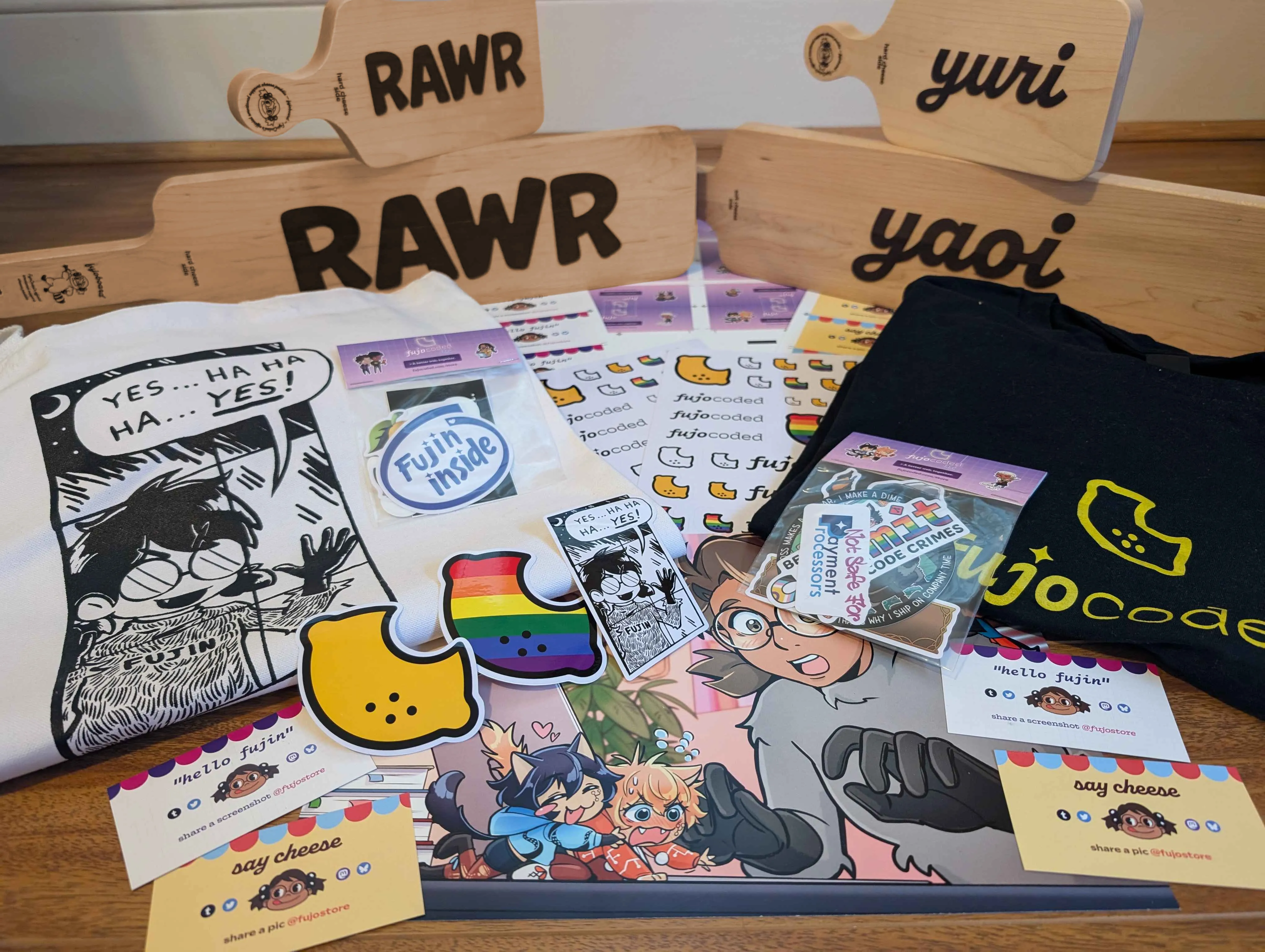 An array of FujoCoded merchandise on a wooden floor. In the back of the array
are four fujoboards, with a chibi version perching on top of the two larger hime
versions, all standing on end so the inscribed text is visible. Two say “rawr”
and the other two say “yuri” and “yaoi.” Underneath the other merchandise are
several sheets of FujoCoded logo stickers and a print of Boba-tan watching Git
and GitHub squabble. On the left and right of the pile are two folded tee
shirts. On top of the tee shirts are sticker packs and large FujoCoded lemon
logo stickers. At the front of the array are four packing cards with FujoCoded
branding that say either “hello fujin” or “say cheese.”